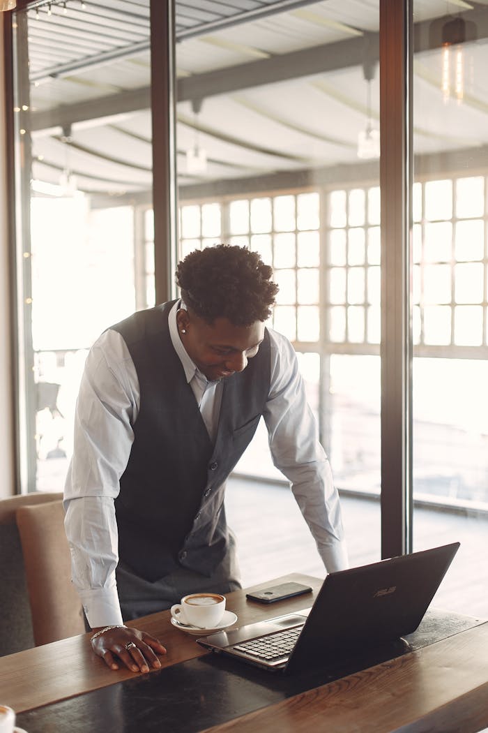 Smiling young African American male entrepreneur in formal clothes working on laptop while standing with hands on table in light modern cafe