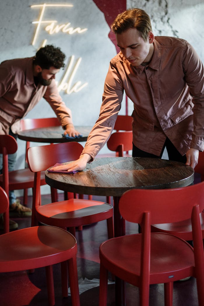 Two staff members prepare a modern cafe interior with red chairs and neon lighting.